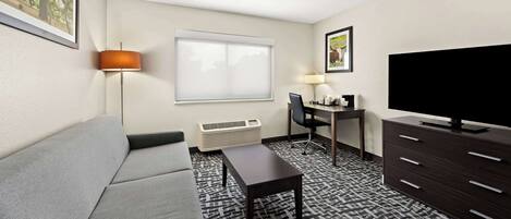 A hotel room with a gray sofa, a small table, a TV, a desk with a chair, and a patterned carpet.