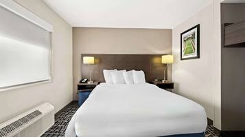 Une chambre d’hôtel avec un grand lit, une table de chevet, une lampe, un tableau encadré au mur et une fenêtre avec des stores.