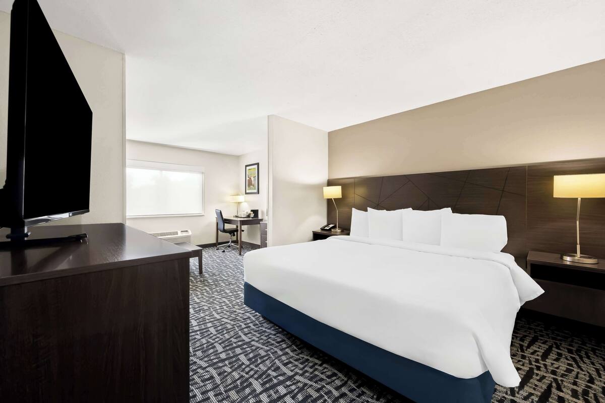 A hotel room with a large bed, a desk, a chair, a TV, and a lamp.