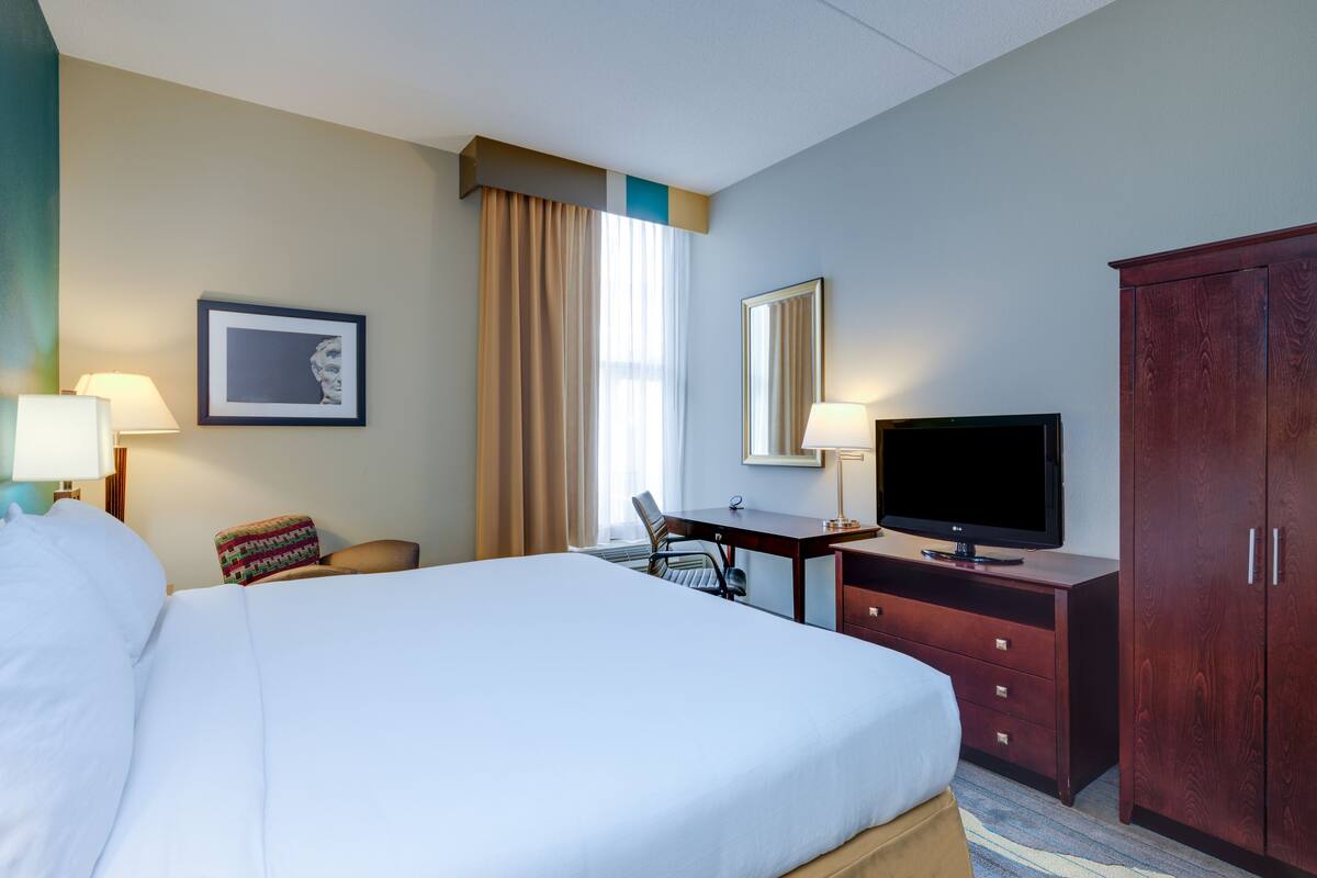 A hotel room with a large bed, a desk with a chair, a television, a wardrobe, and a window with curtains.