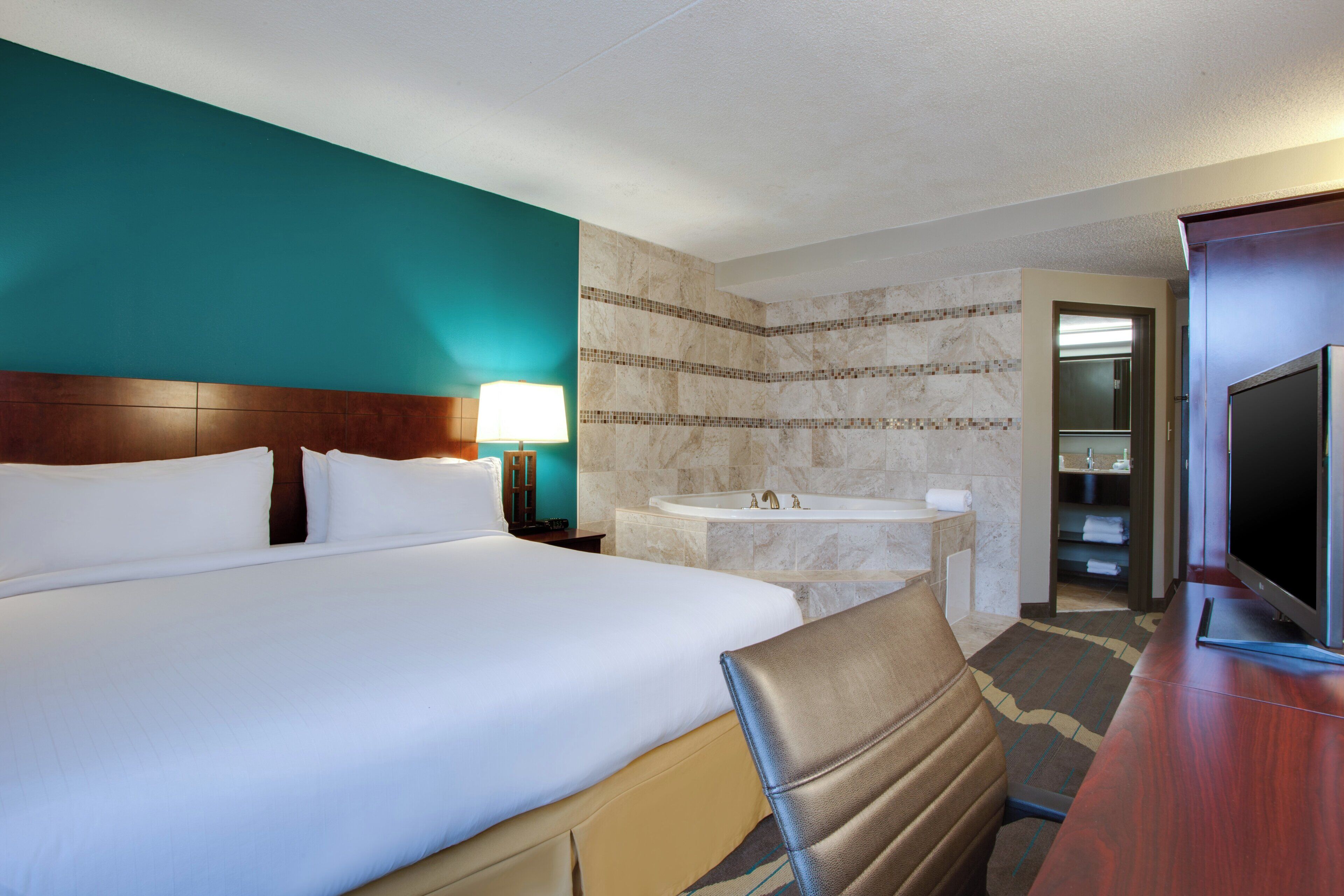 A hotel room with a large bed, a bedside table with a lamp, a bathroom with a sink and mirror, and a television on a wooden table.