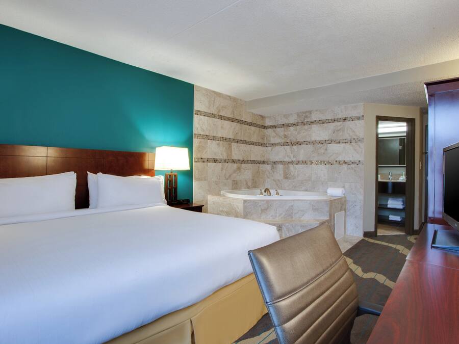 A hotel room with a large bed, a bedside table with a lamp, a bathroom with a sink and mirror, and a television on a wooden table.