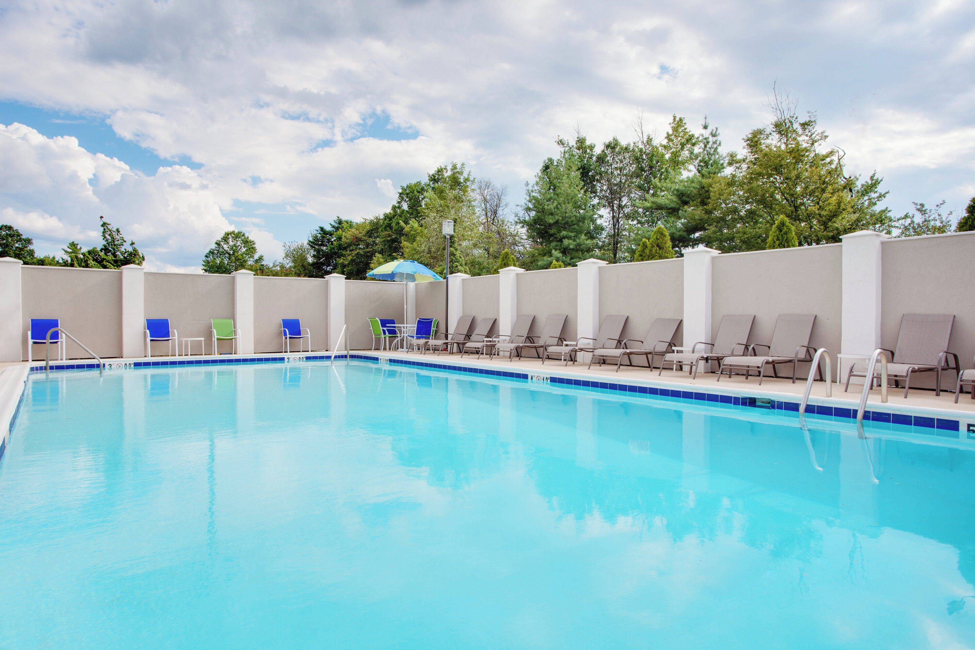 A swimming pool with lounge chairs and a covered area, surrounded by a low wall and trees.
