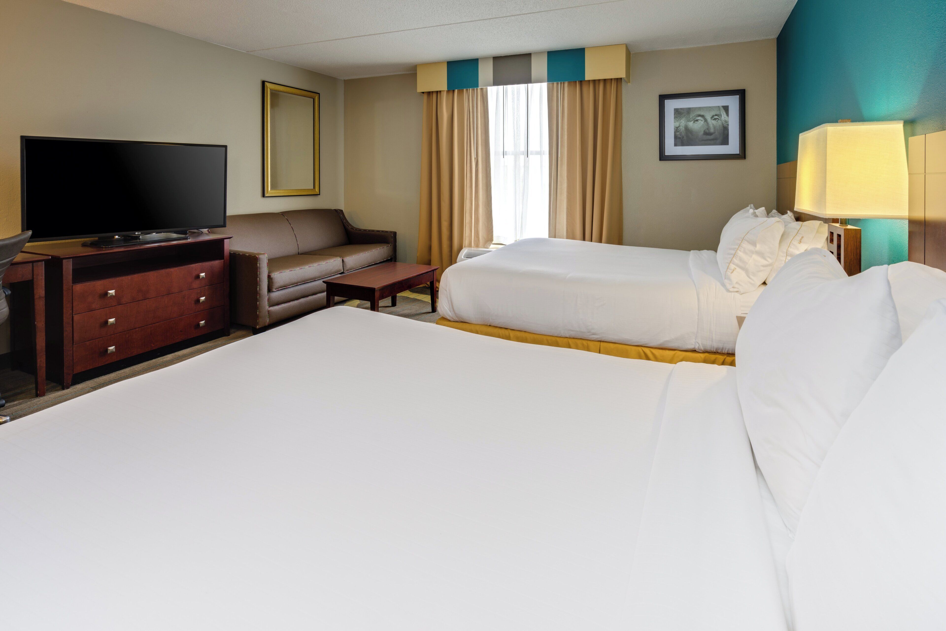 A hotel room with a large bed, a smaller bed, a sofa, a TV, a desk, and a lamp.