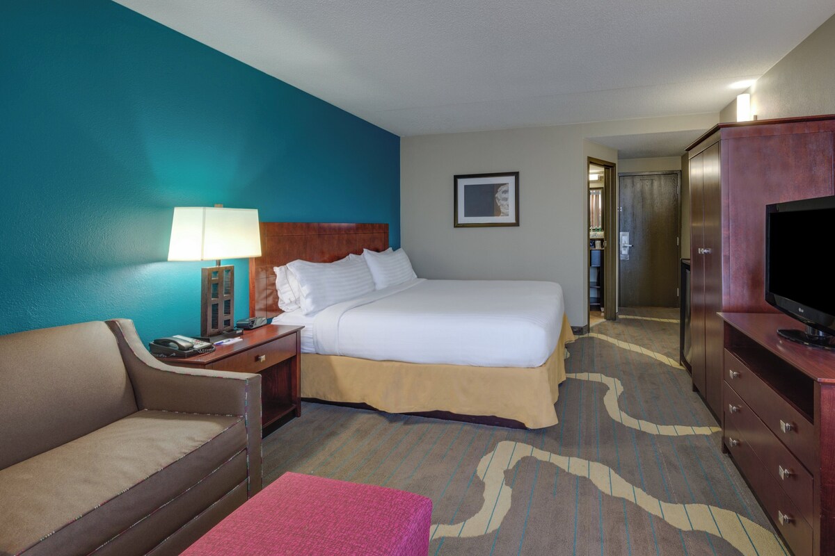 A hotel room with a bed, a desk with a lamp, a television, a sofa, and a carpet with a pattern.