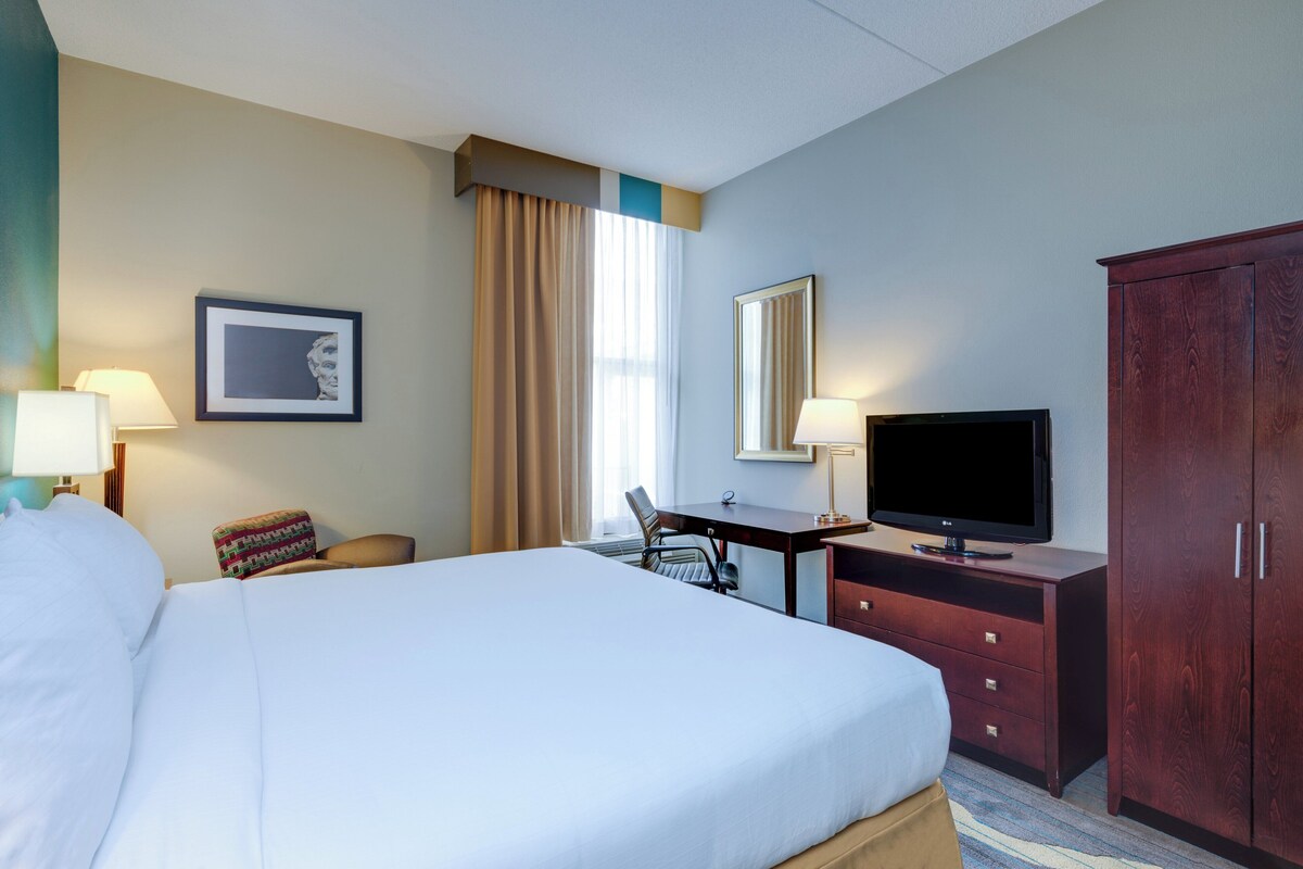A hotel room with a large bed, a desk with a chair, a television, a wardrobe, and a window with curtains.