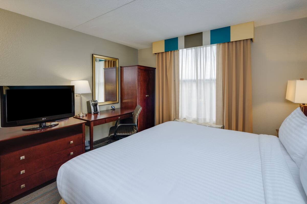 A hotel room with a large bed, a desk with a television, a chair, a wardrobe, and a window with curtains.