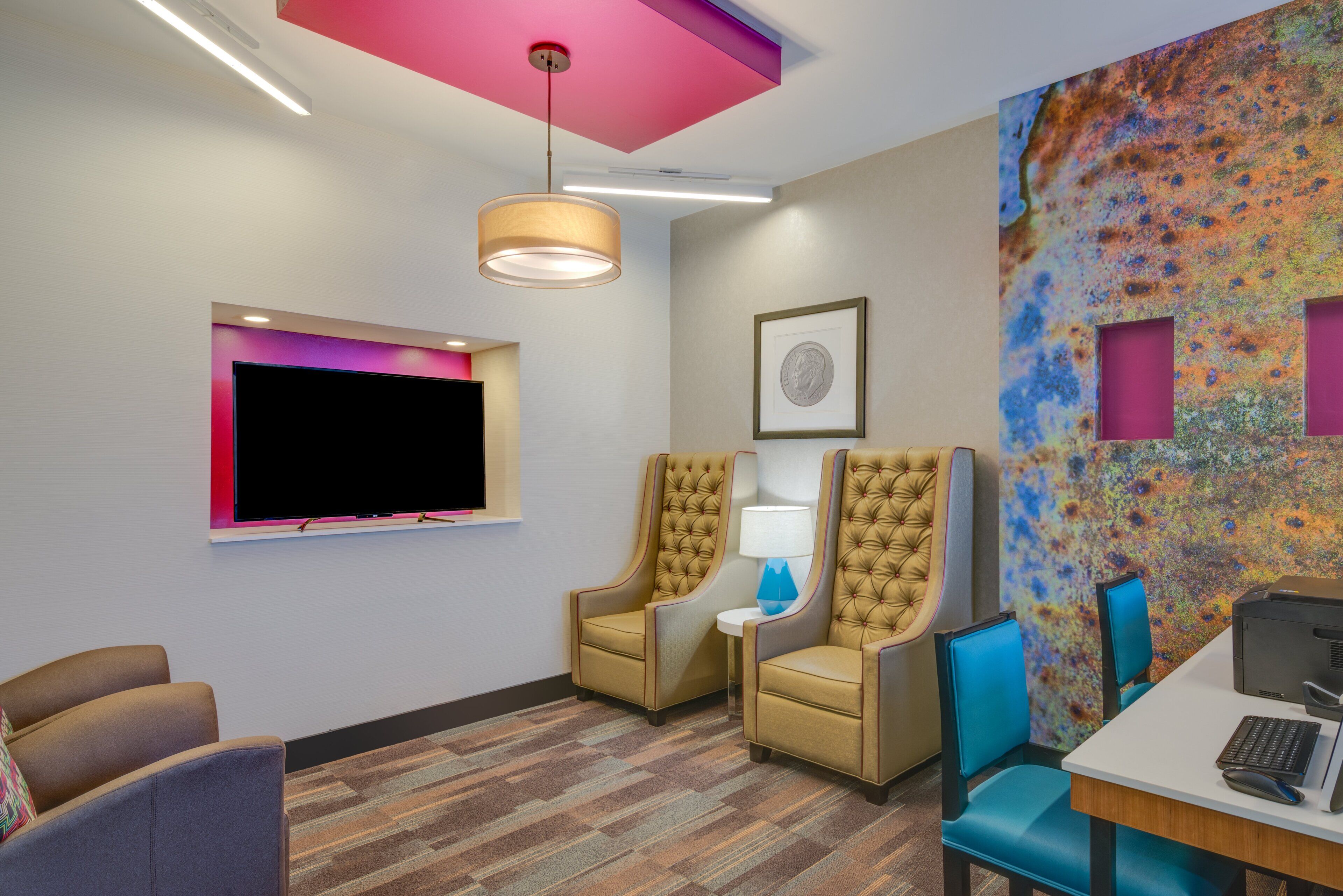 A modern room with a flat-screen TV, two tufted chairs, a desk with a computer, and a colorful abstract wall art.