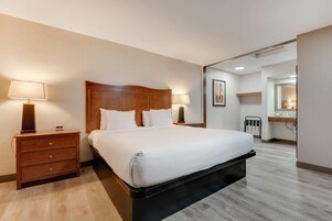 Habitación de hotel con una cama grande, cabecera de madera, dos mesitas de noche con lámparas, un pequeño aparador de madera y un baño visible a través de una puerta abierta.