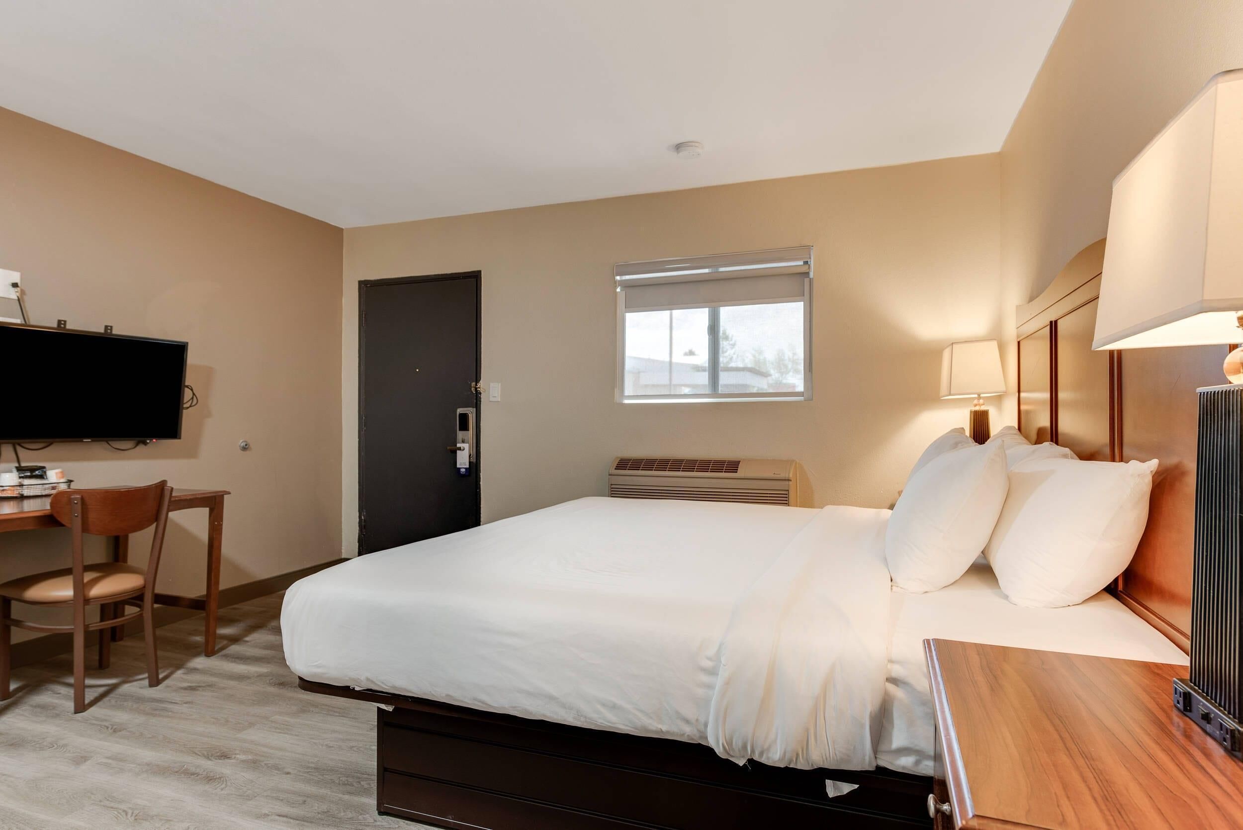 A hotel room with a bed, a desk with a chair, a television, a window with blinds, and a wall-mounted lamp.