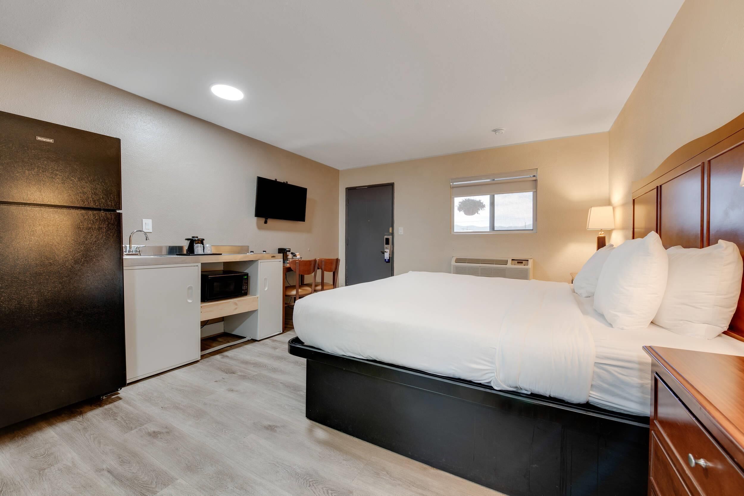A compact hotel room with a kitchenette, a bed with white linens, a flat-screen TV, and a window with a view.