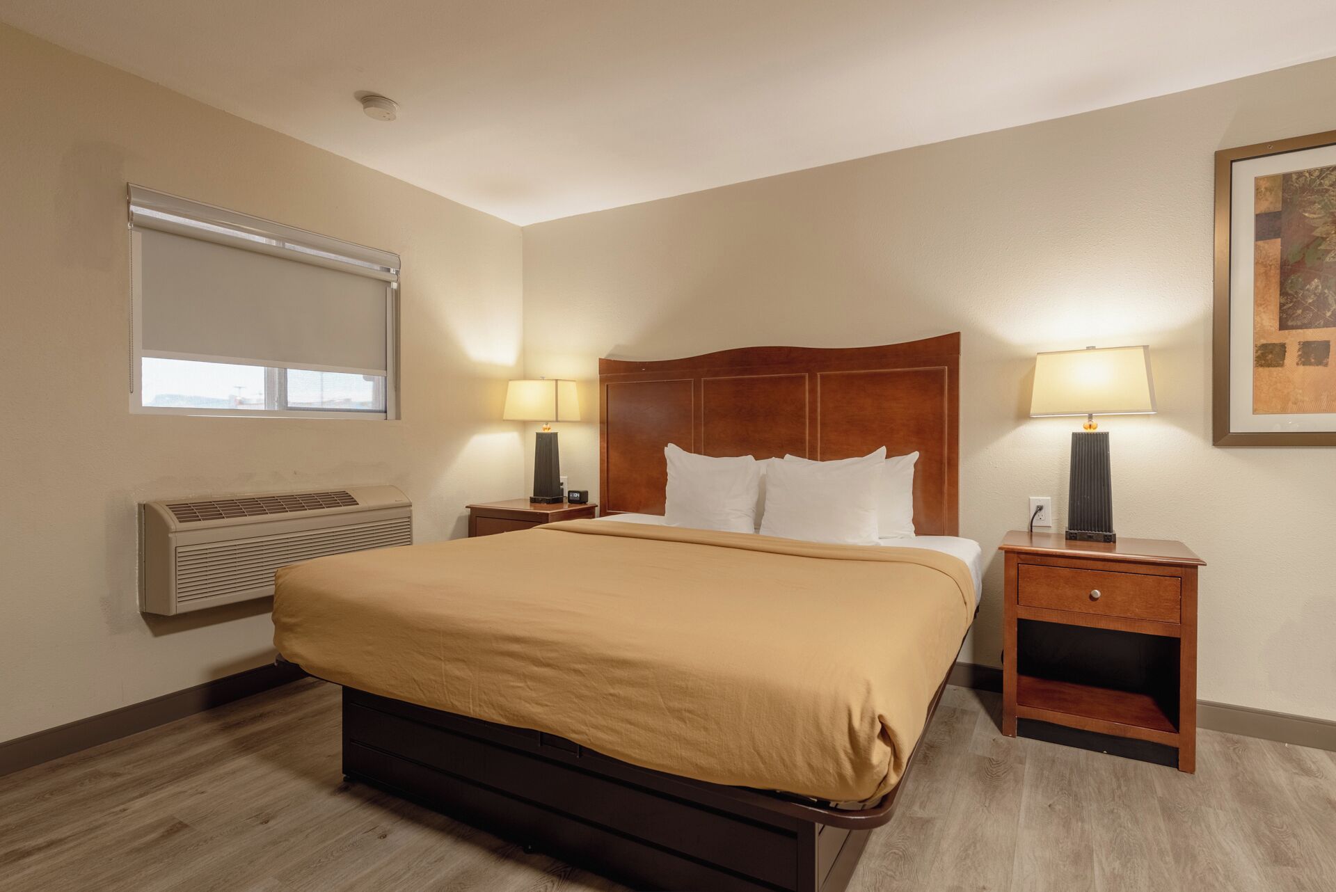 Una habitación de hotel con una cama grande, dos mesitas de noche con lámparas, un aire acondicionado de pared y una ventana con persianas.