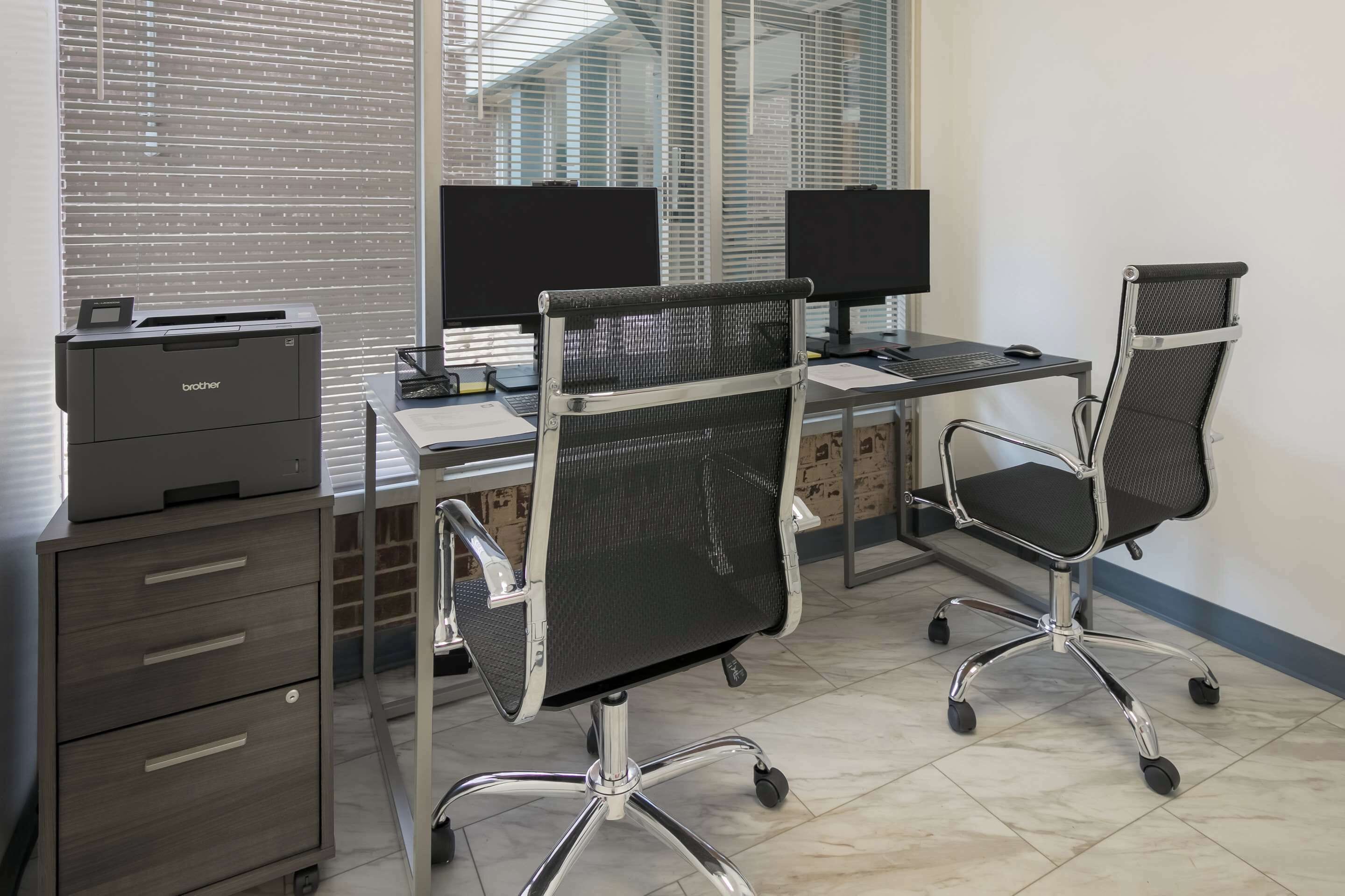 Un bureau moderne équipé de deux postes de travail, chacun comprenant un écran d’ordinateur, des chaises de bureau noires à dossier en maille et une imprimante Brother posée sur un meuble.