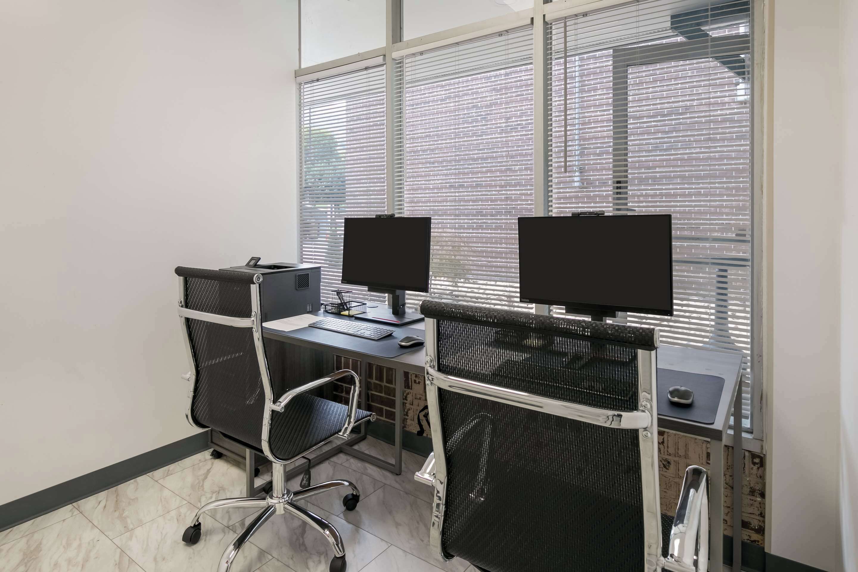 Un espace de bureau moderne comprenant deux bureaux, chacun équipé d’un écran d’ordinateur, d’un clavier et d’une souris. On y trouve également deux chaises de bureau, dont une à roulettes et une autre avec un dossier en tissu ajouré. La pièce est dotée de grandes fenêtres équipées de stores et le sol est carrelé.