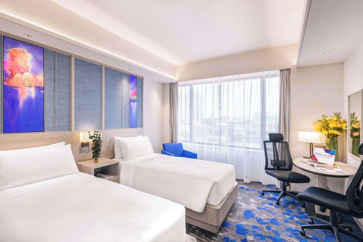 A hotel room with two beds, a desk with a chair, a large window with curtains, and a wall-mounted TV.