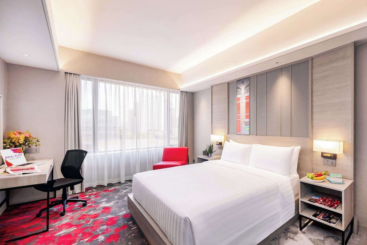 A hotel room with a large bed, a desk with a chair, a red chair, a nightstand with a lamp, and a wall-mounted TV.