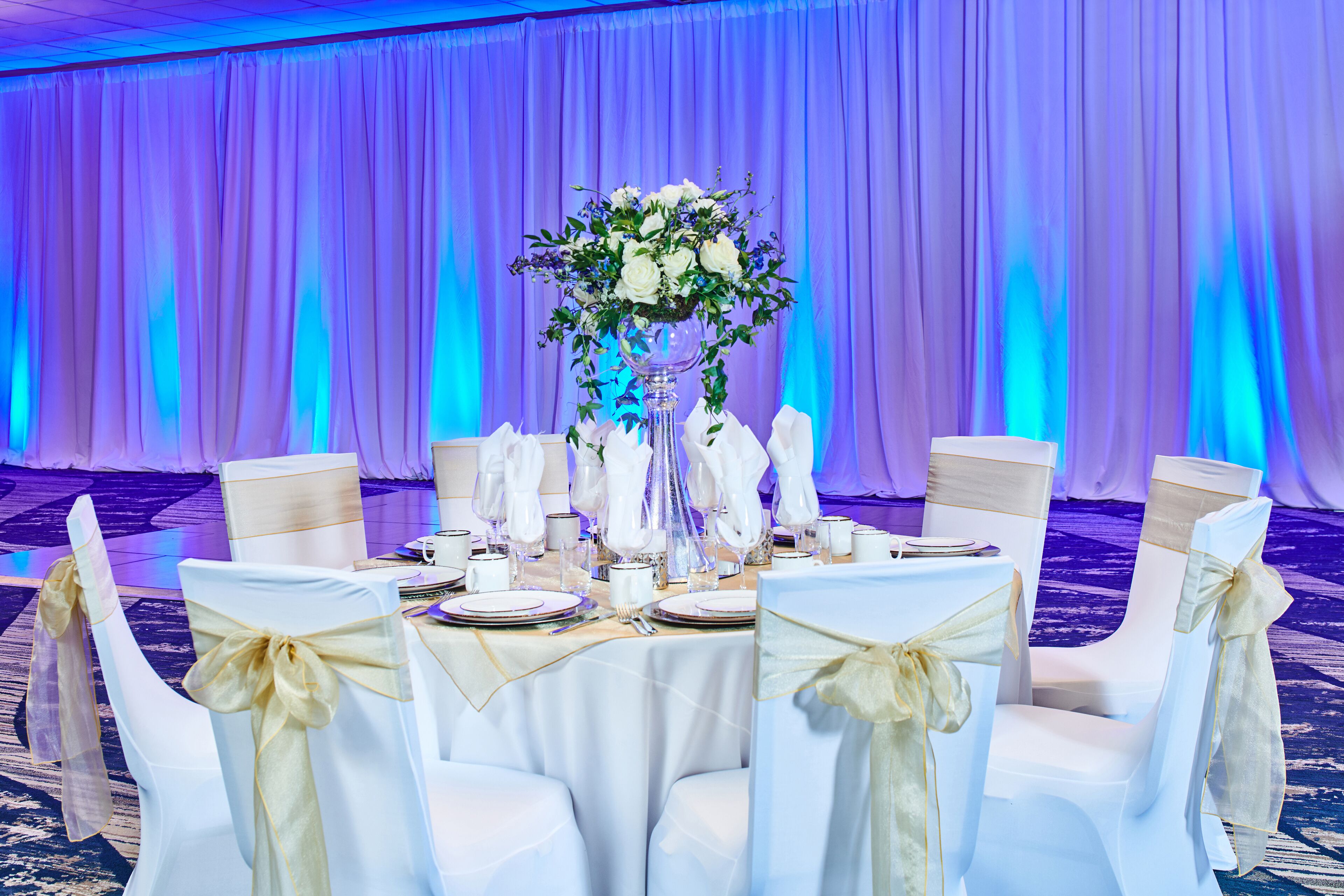 Ein festlich gedeckter Bankettisch mit weißen Tischdecken, goldenen Bändern und einer floralen Tischdekoration.