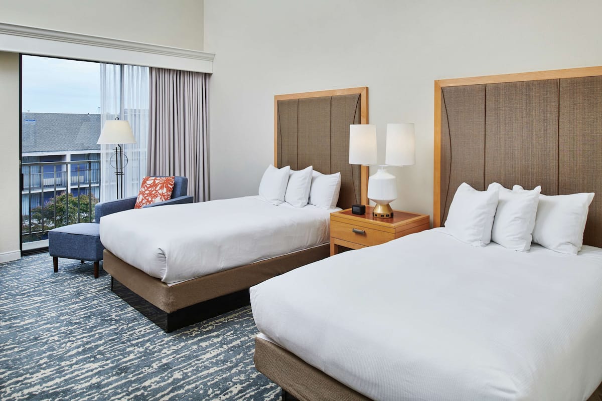 A hotel room with two beds, a chair, a nightstand, and a large window with curtains.