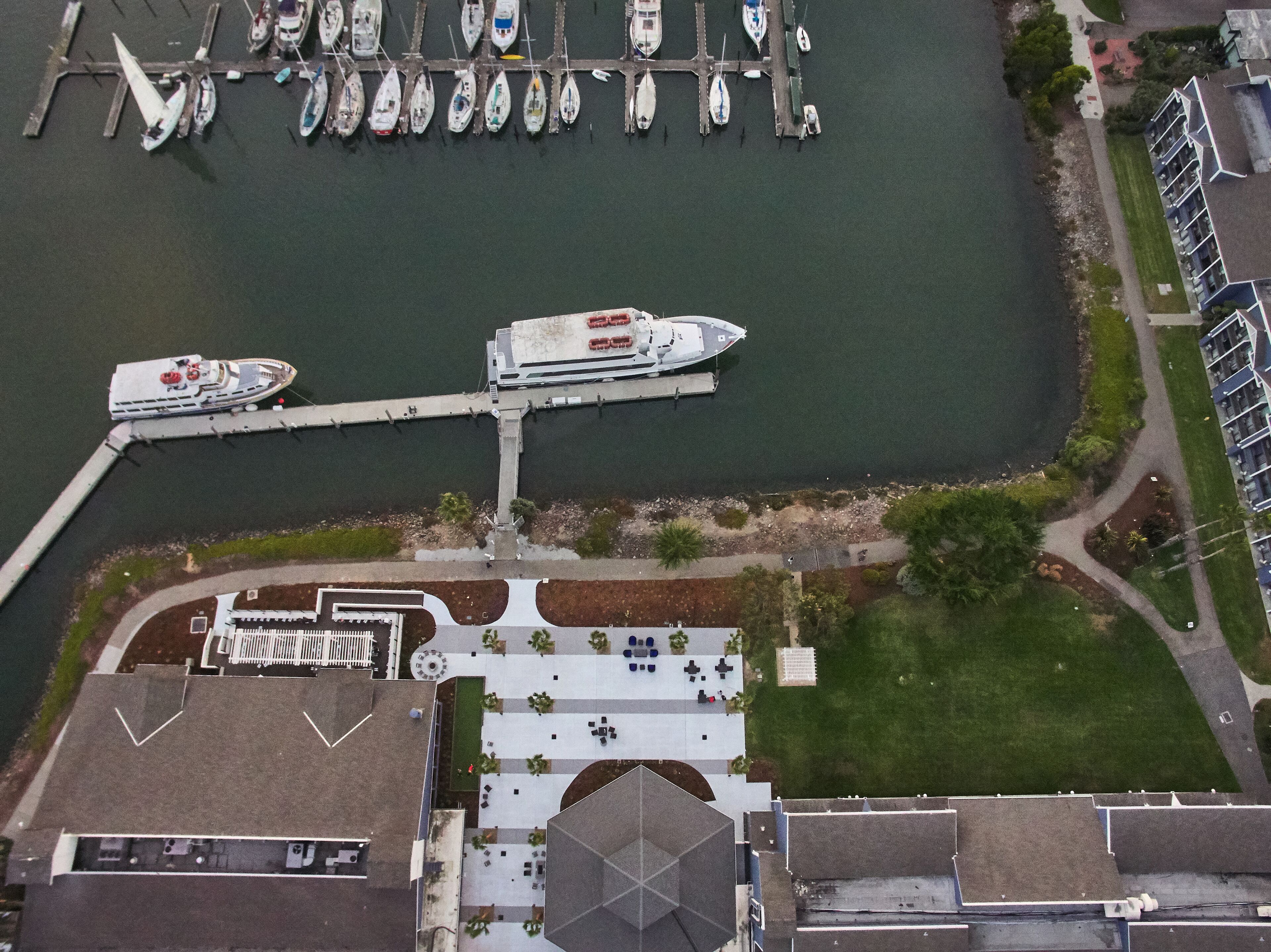 An aerial view of a marina with several boats docked, a pedestrian walkway, and a building with a distinct roofline.