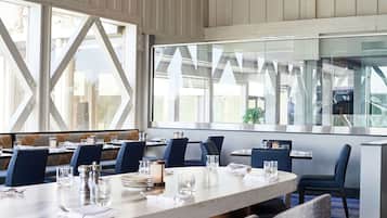 Un comedor moderno con ventanas amplias, sillas tapizadas en azul y mesas ordenadas con copas y cubiertos.