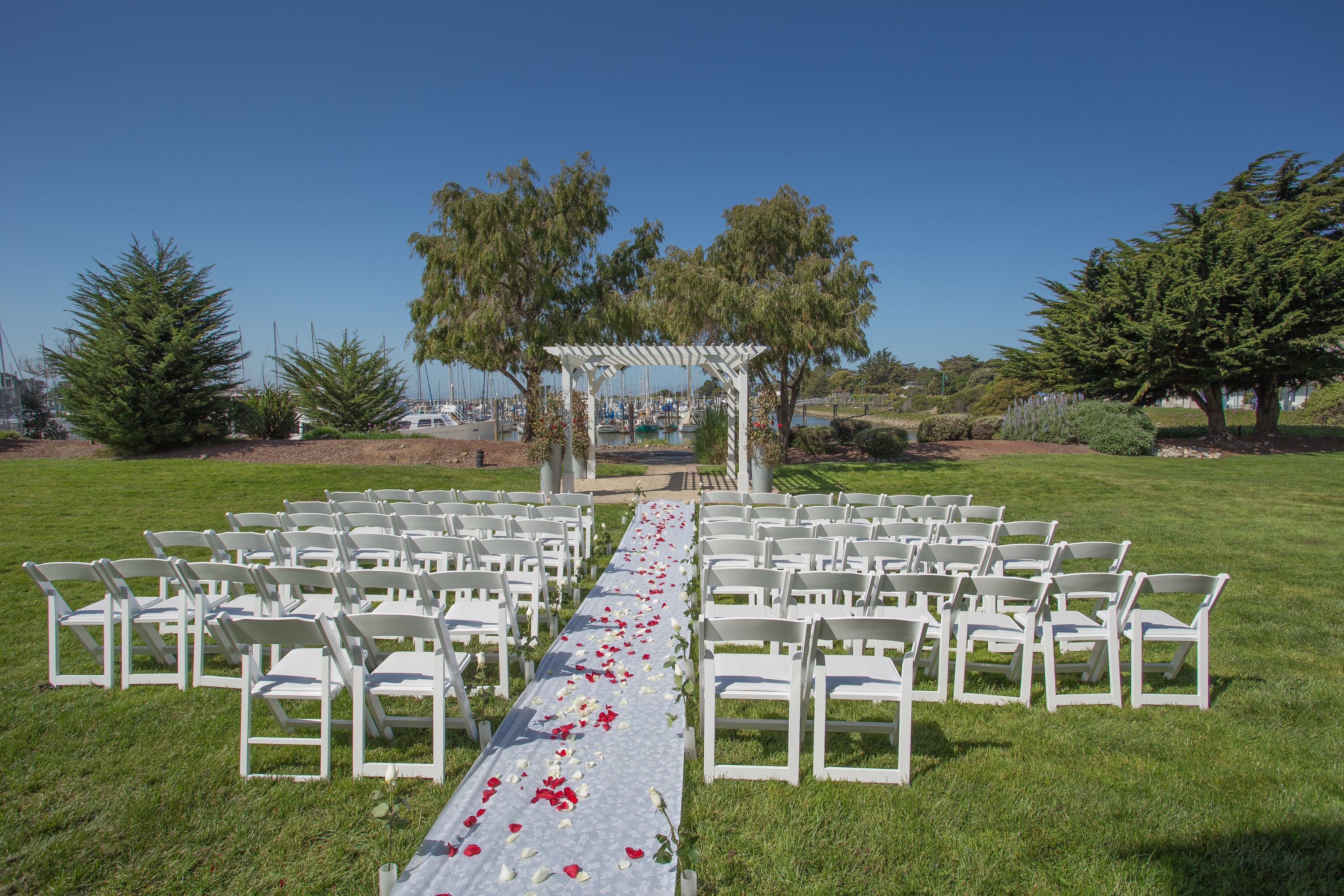An outdoor wedding ceremony setup with white chairs arranged in rows, a white aisle with red flower petals, and a white pergola structure in the background.