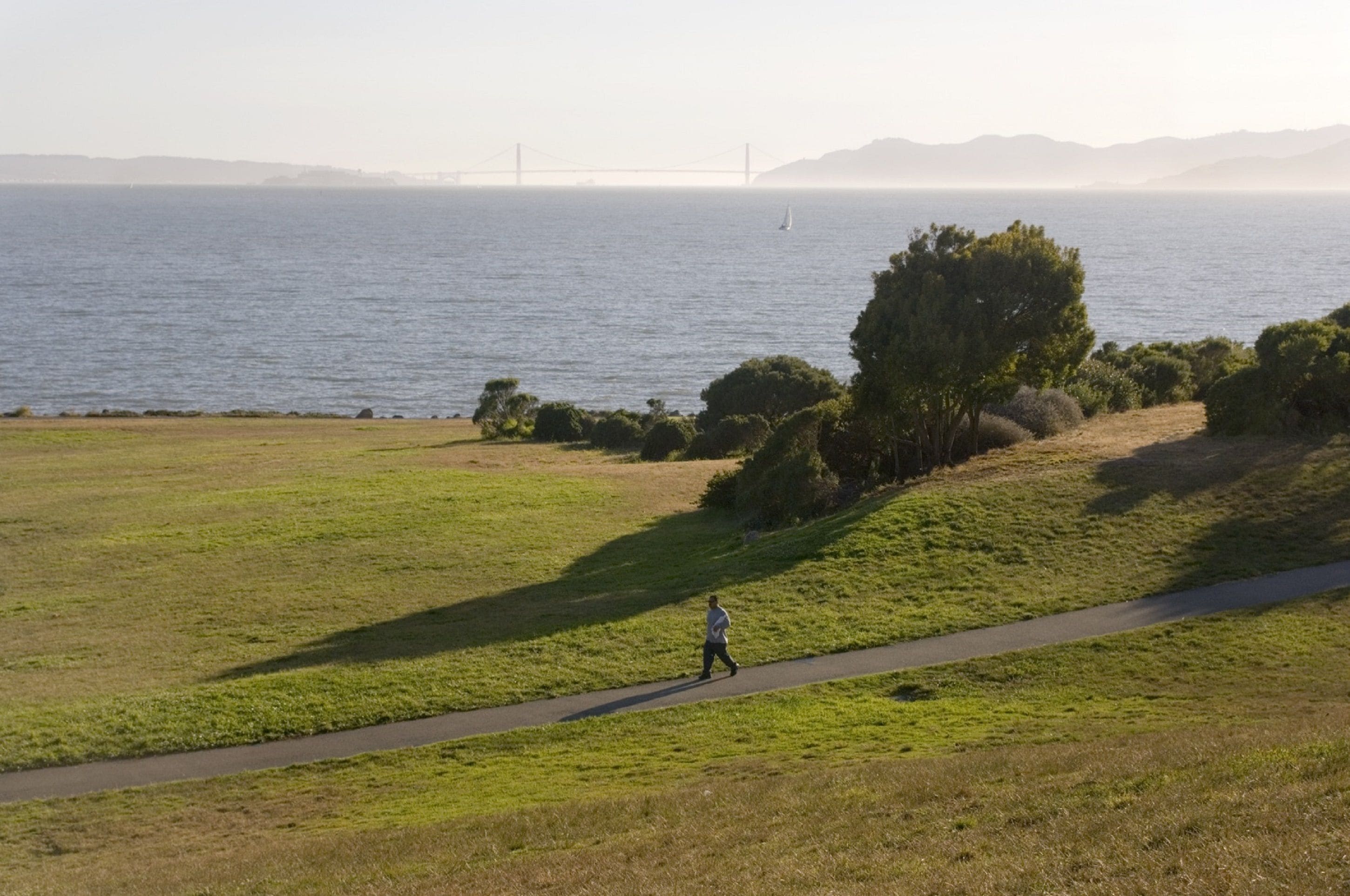 A person walking on a path beside a body of water with a bridge in the background.