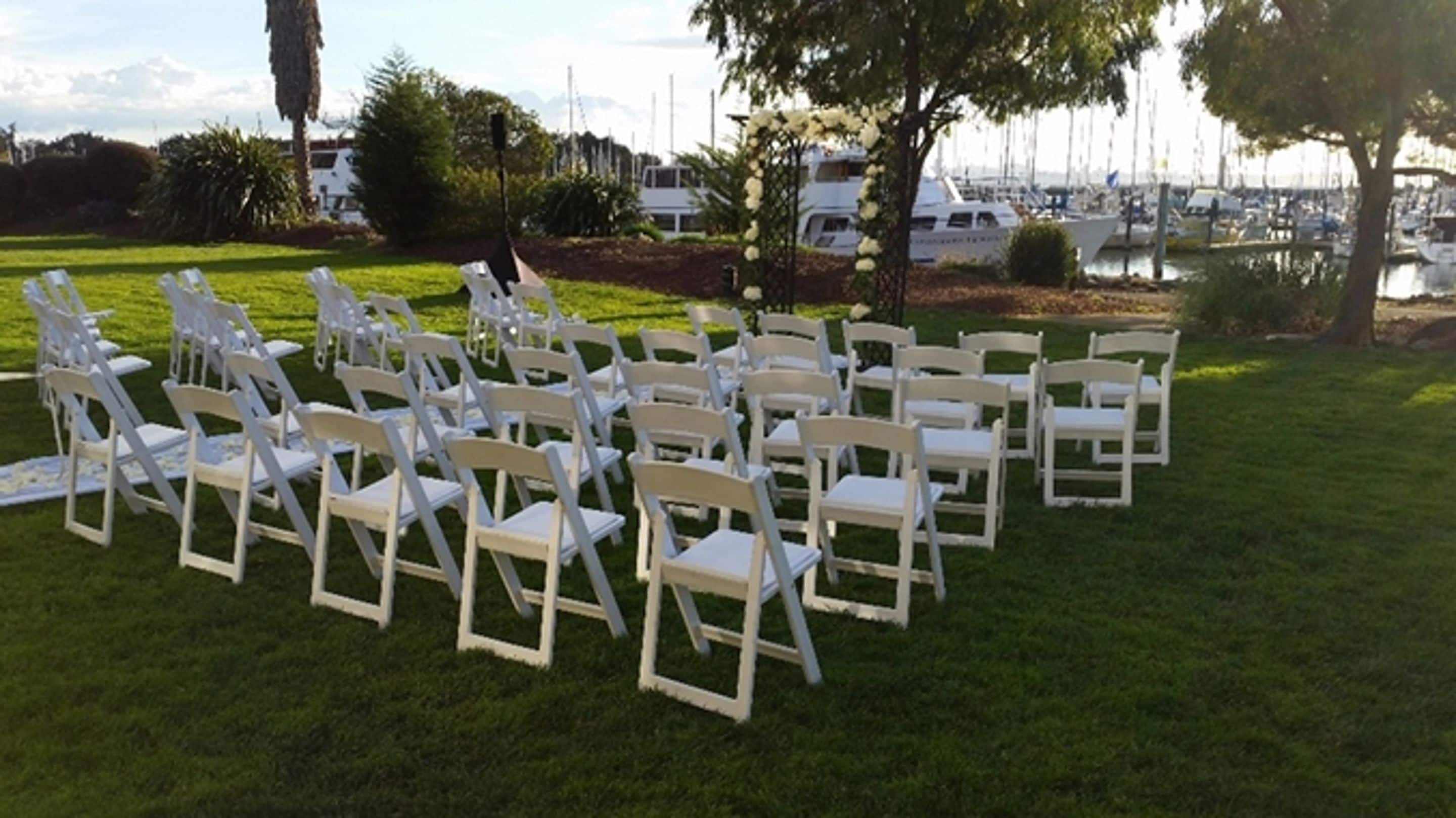 Rows of white folding chairs arranged on a grassy lawn with a marina and boats in the background.