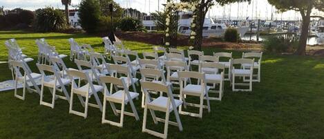 Rows of white folding chairs arranged on a grassy lawn with a marina and boats in the background.