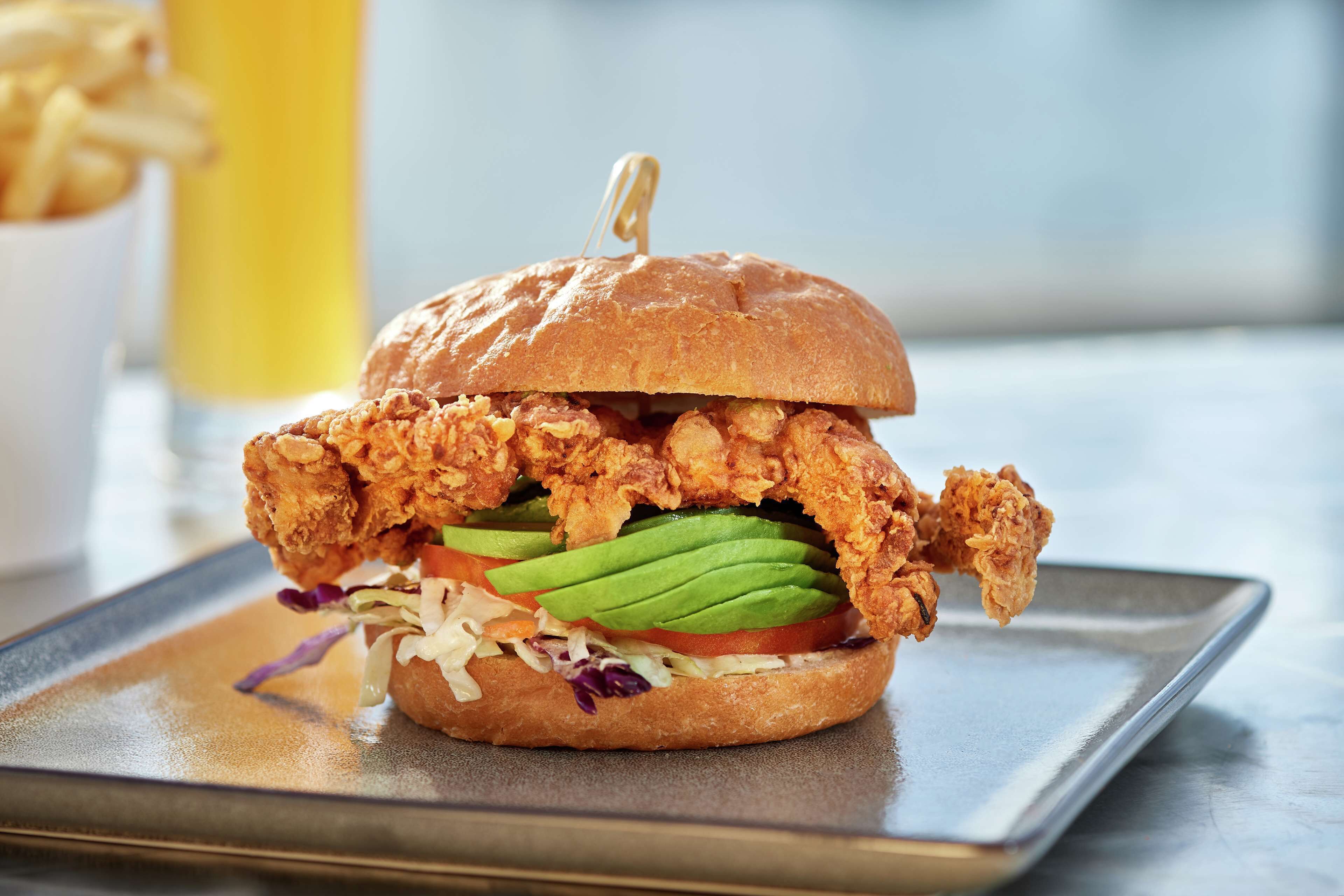 A fried chicken sandwich with a bun, avocado, tomato, and lettuce on a metal tray.