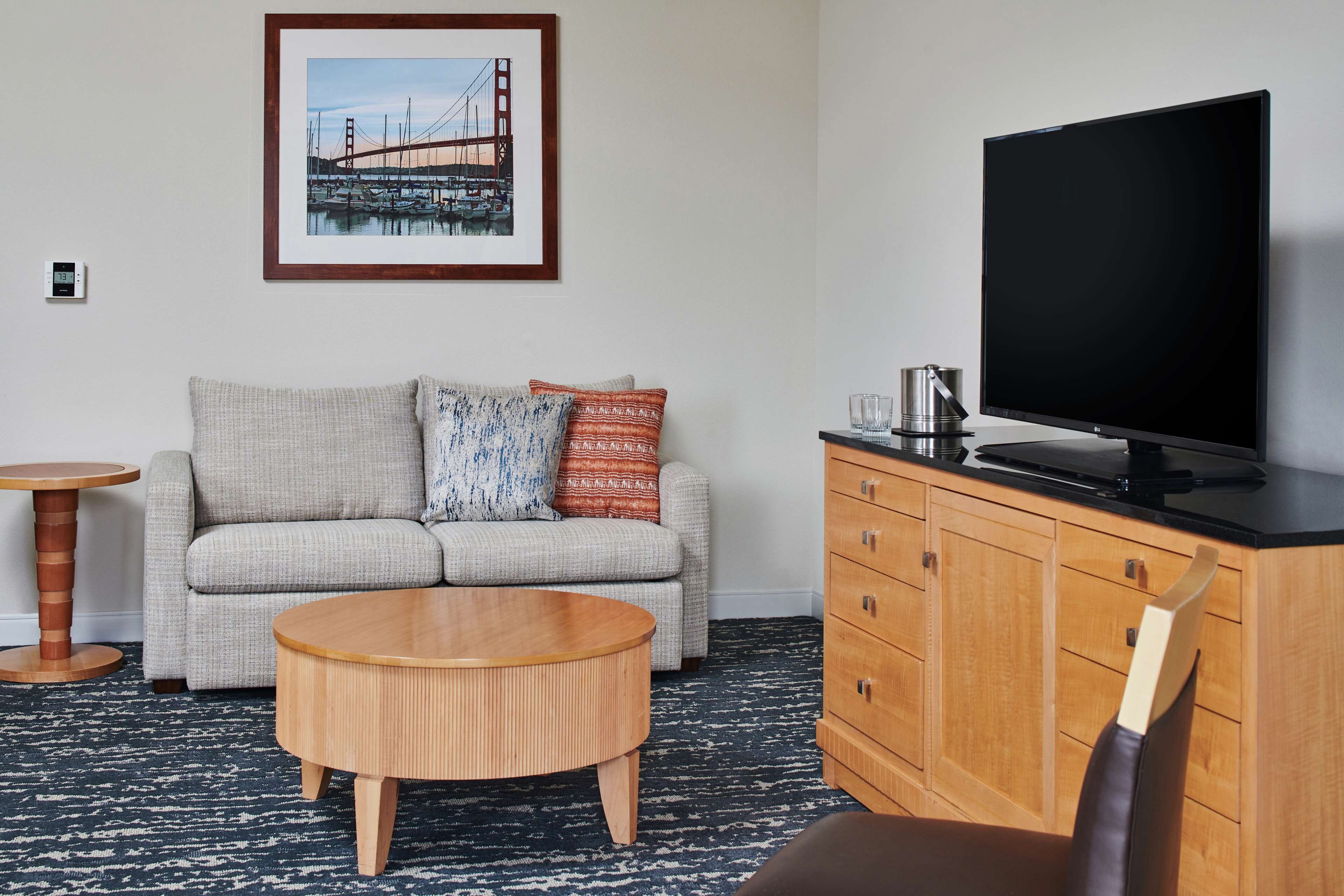 A hotel room with a sofa, a round wooden coffee table, a television on a wooden cabinet, and a framed picture of a bridge on the wall.