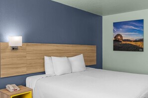 Habitación de hotel con cabecera de madera, cama con ropa de cama blanca, lámpara de noche, teléfono y un cuadro de paisaje en la pared.