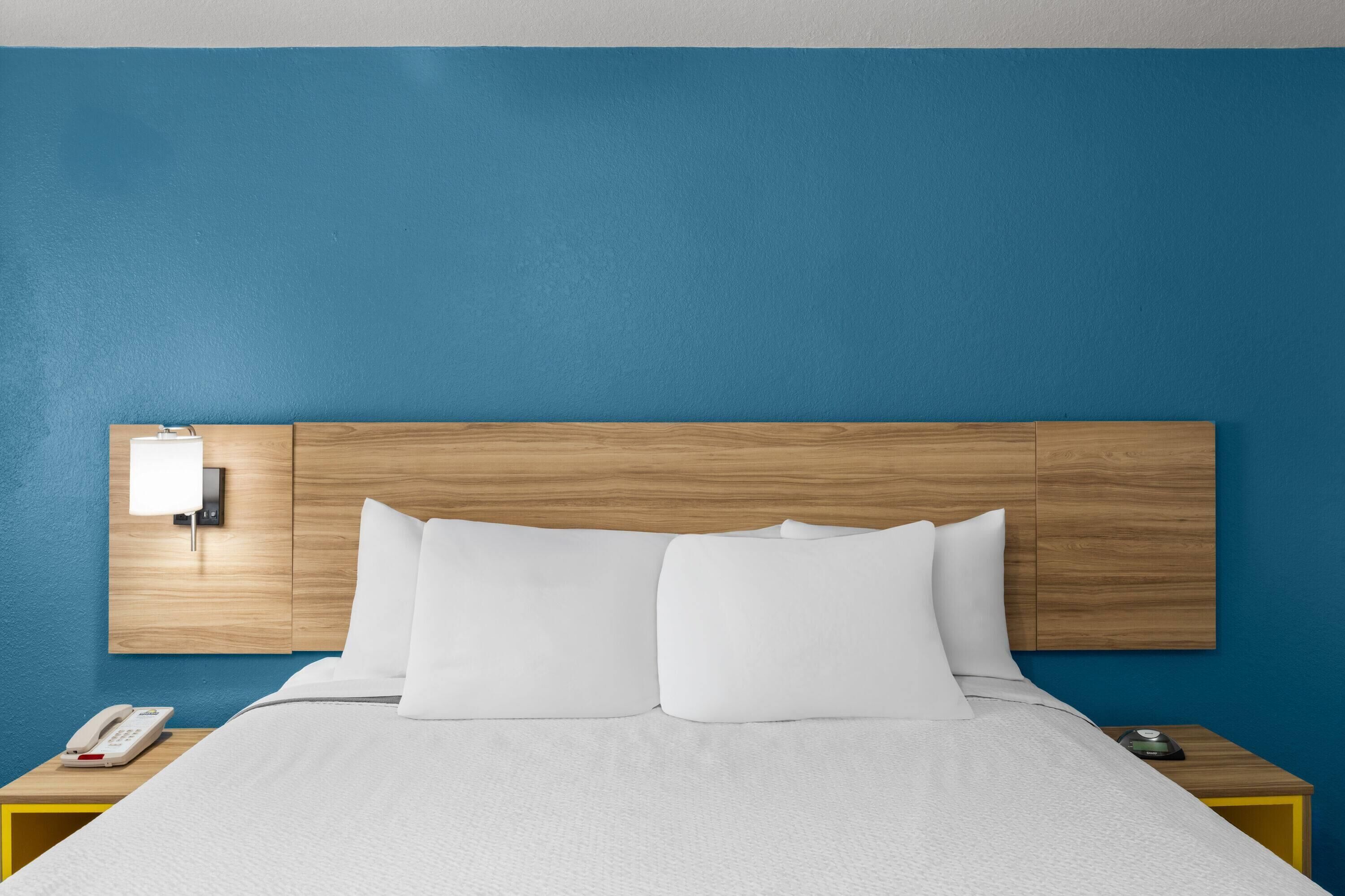 Habitación de hotel con cabecera de madera, cama blanca, mesita de noche con teléfono y luz de pared.