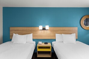 A hotel room with a wooden headboard, two beds, white bedding, a bedside table with a lamp, and a round mirror on the wall.