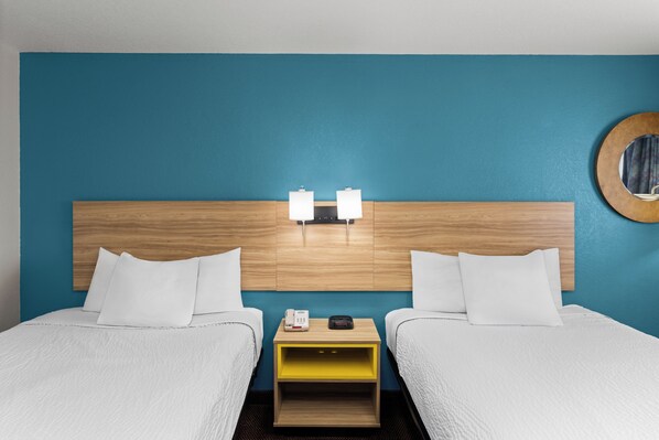 A hotel room with a wooden headboard, two beds, white bedding, a bedside table with a lamp, and a round mirror on the wall.