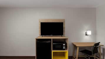 A hotel room with a TV on a wooden stand, a microwave, a small yellow shelf, and a desk with a chair.
