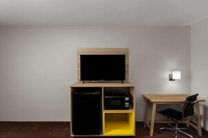 A hotel room with a TV on a wooden stand, a microwave, a small yellow shelf, and a desk with a chair.