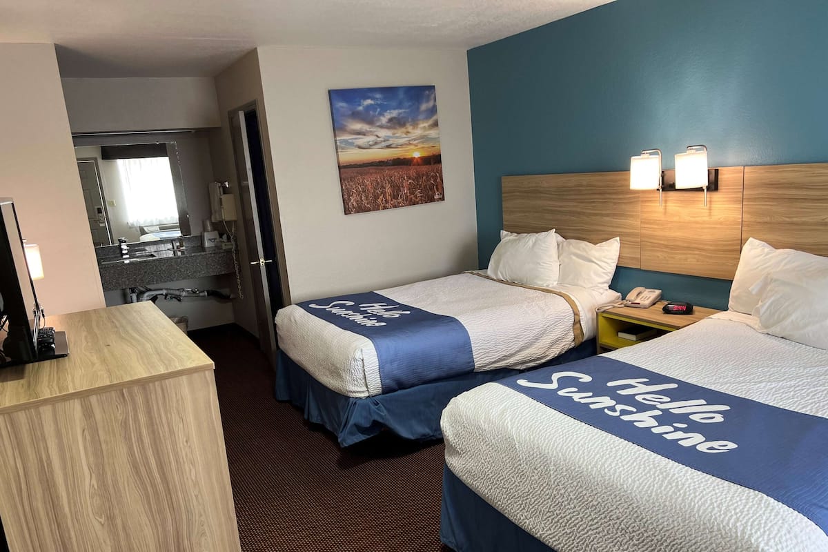 A hotel room with two beds, a desk, a mirror, a sink, and a painting of a sunset.
