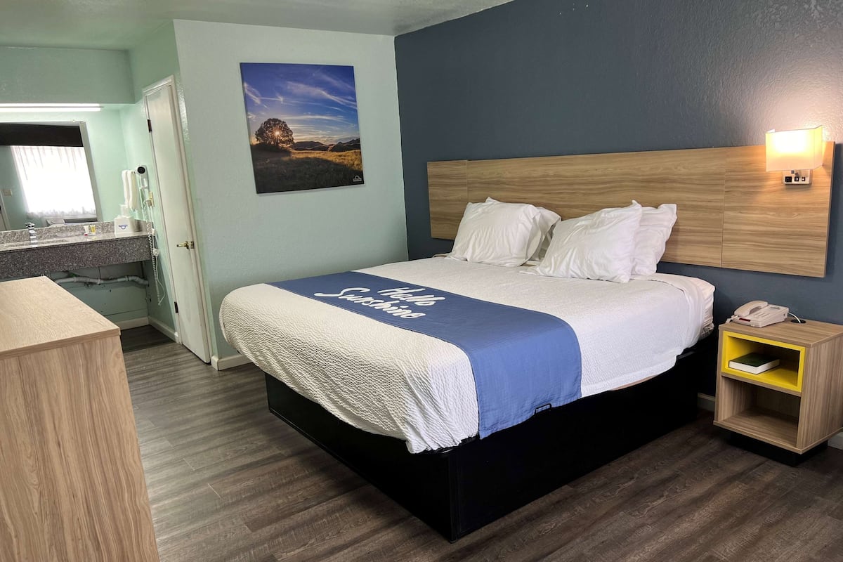 A hotel room with a bed, a bedside table with a lamp, a nightstand, a bathroom with a sink and mirror, and a painting on the wall.