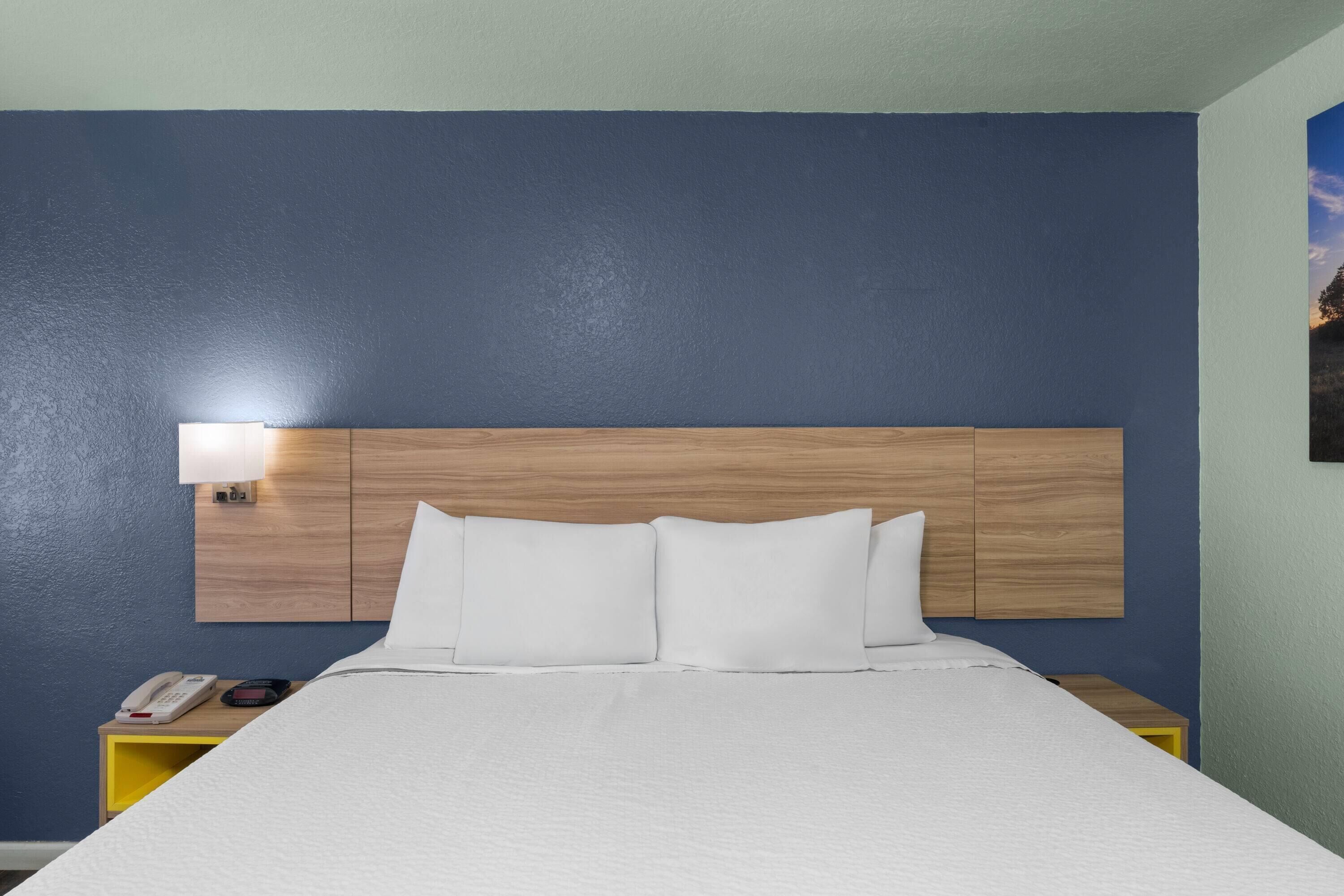 A hotel room with a wooden headboard, a white bedspread, and a bedside table with a lamp and a phone.