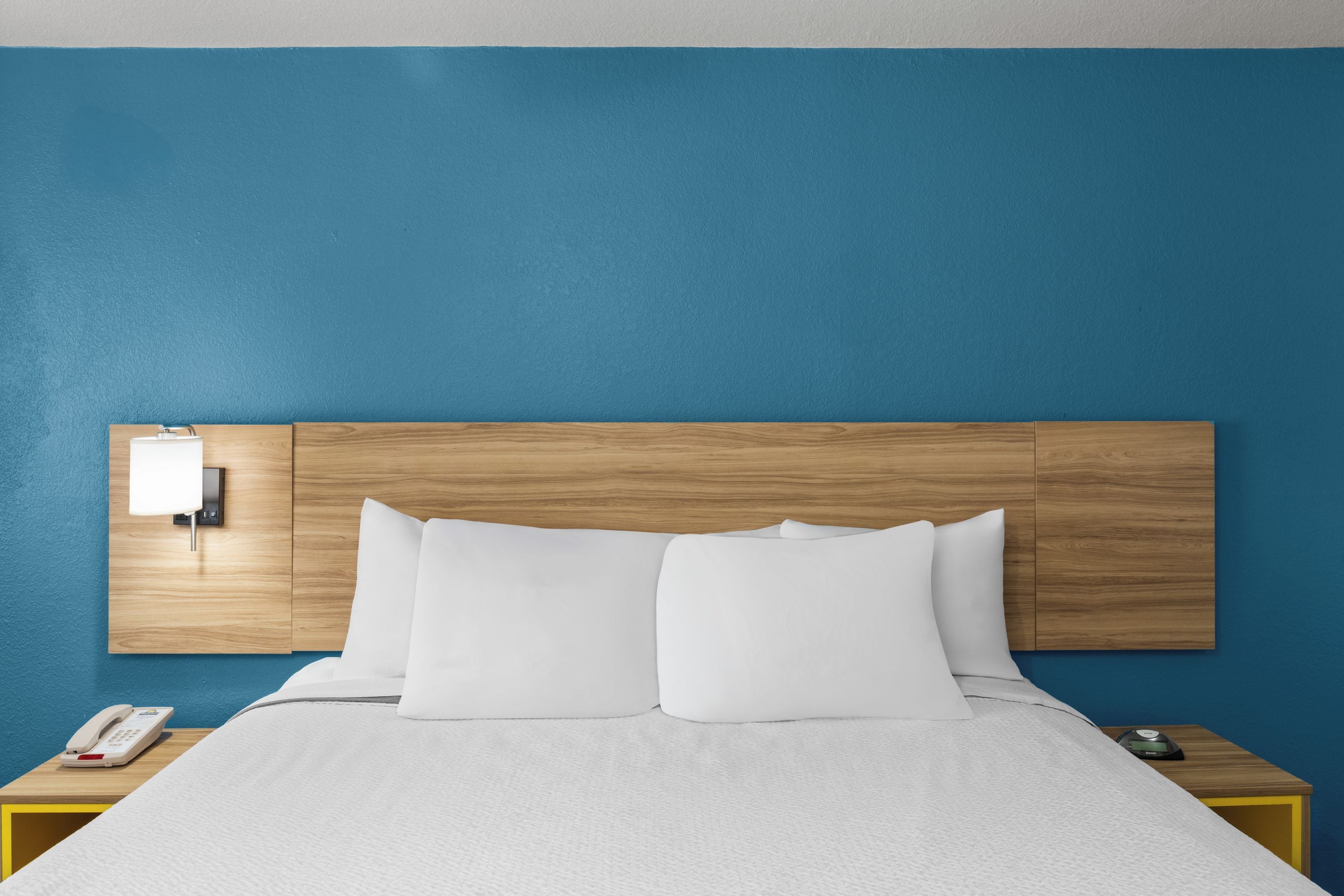 A hotel room with a wooden headboard, white bedding, and a bedside table with a phone and a digital clock.