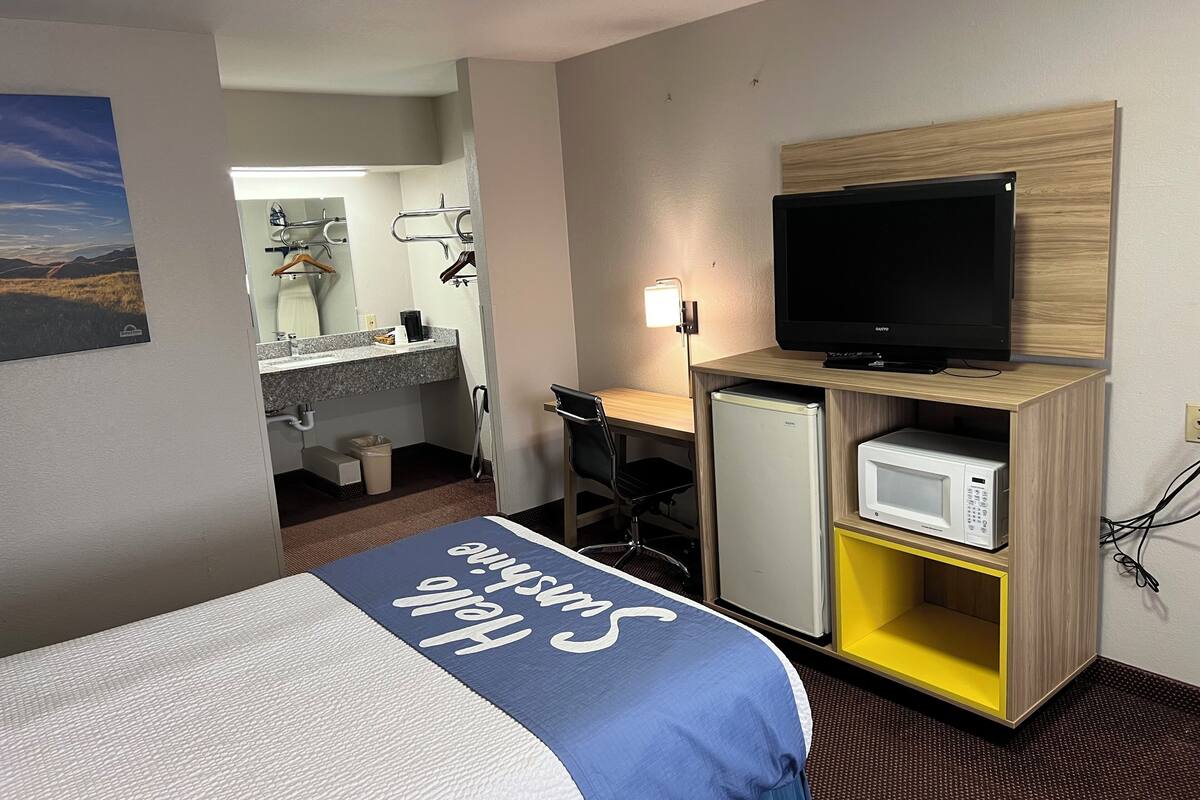 Una habitación de hotel con una cama, un escritorio con computadora, televisión, microondas y refrigerador pequeño.