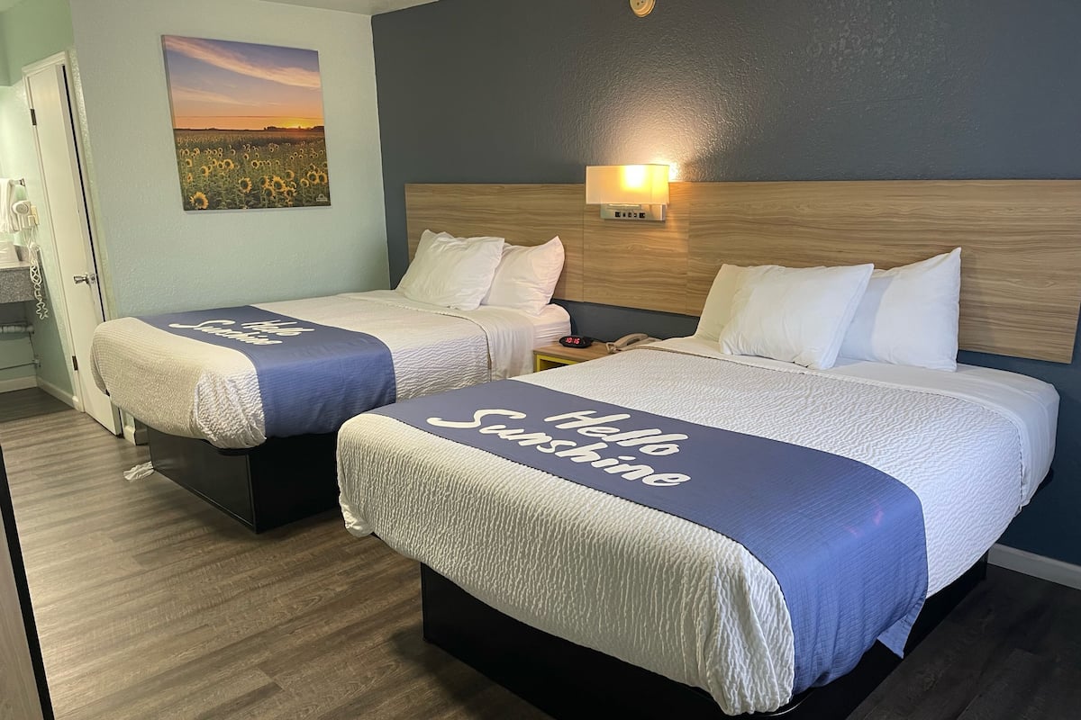 A hotel room with two beds, each with a 'Hello Sunshine' bedspread, a nightstand with a lamp, and a painting of a sunset on the wall.