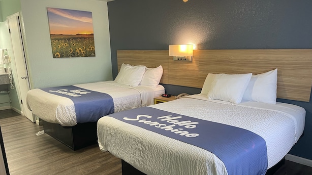 Una habitación de hotel con dos camas, cada una con colcha 'Hello Sunshine', una mesita de noche con lámpara y un cuadro de un atardecer en la pared.