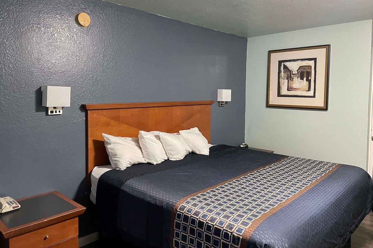 A hotel room with a wooden headboard, a bed with a patterned bedspread, a bedside table with a phone, and a wall-mounted light fixture.
