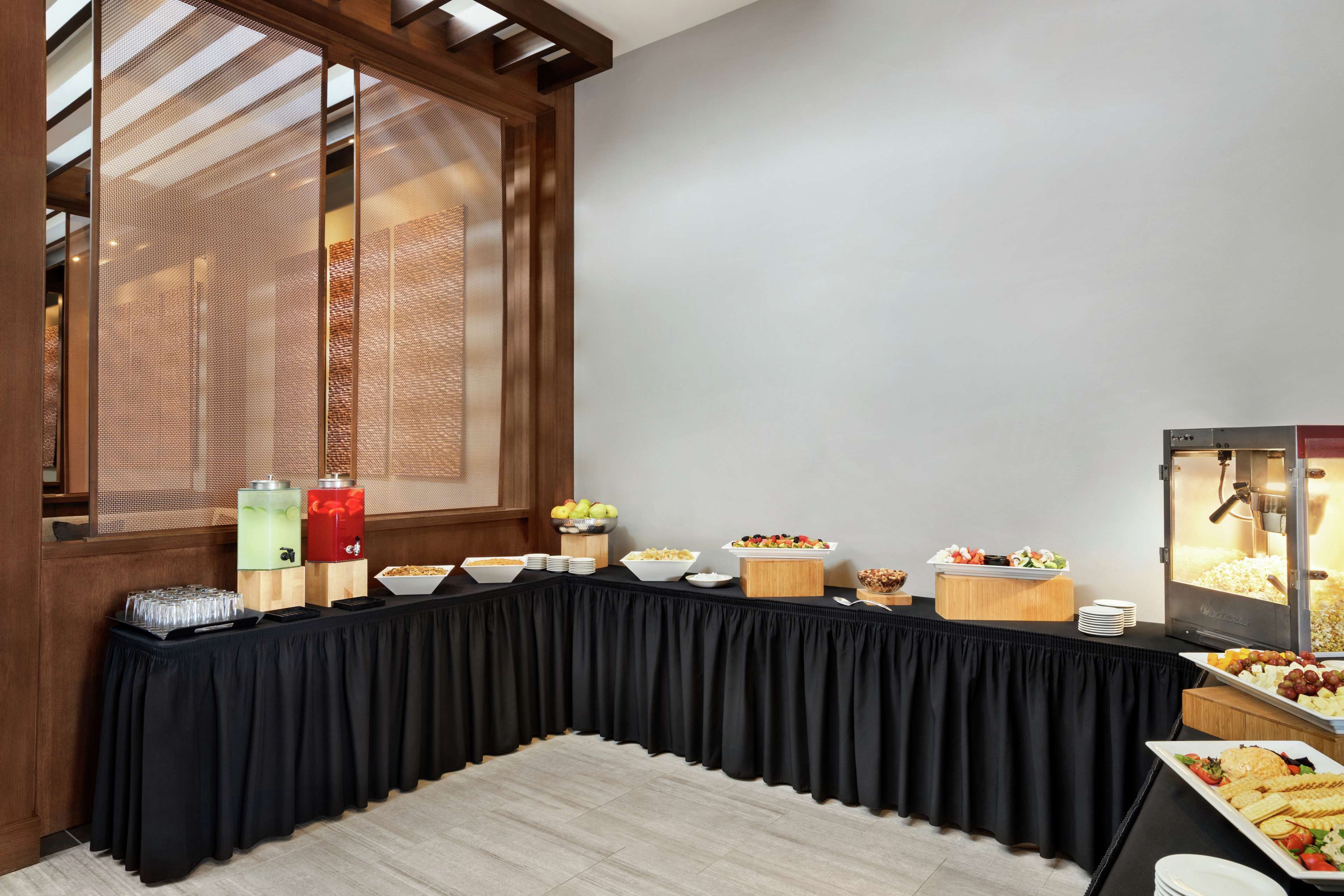 A buffet setup with various food items and beverages, a large window with blinds, and a modern interior design.