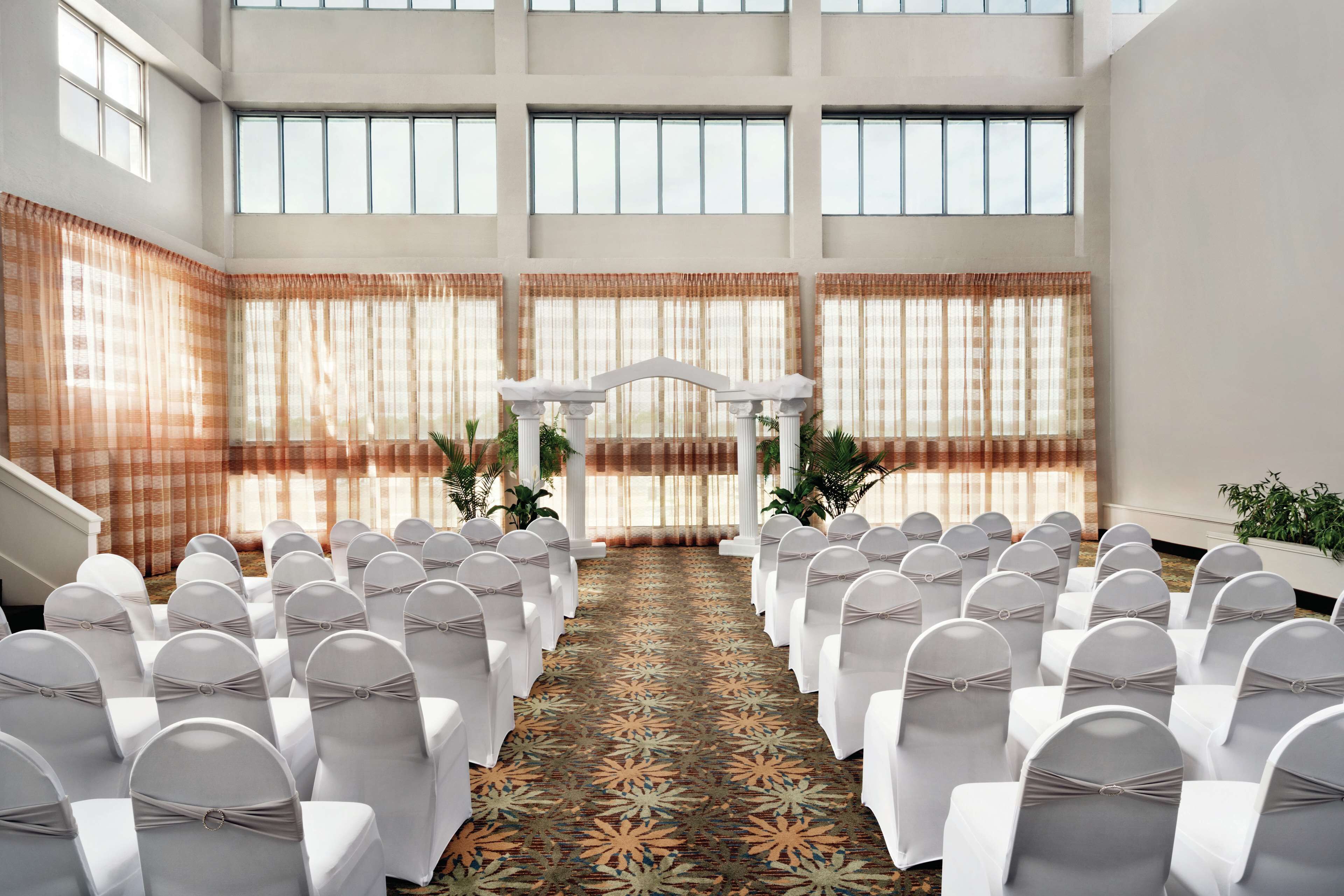 Un salón de conferencias con filas de sillas blancas, una alfombra estampada y un pasillo central que conduce a un arco blanco.