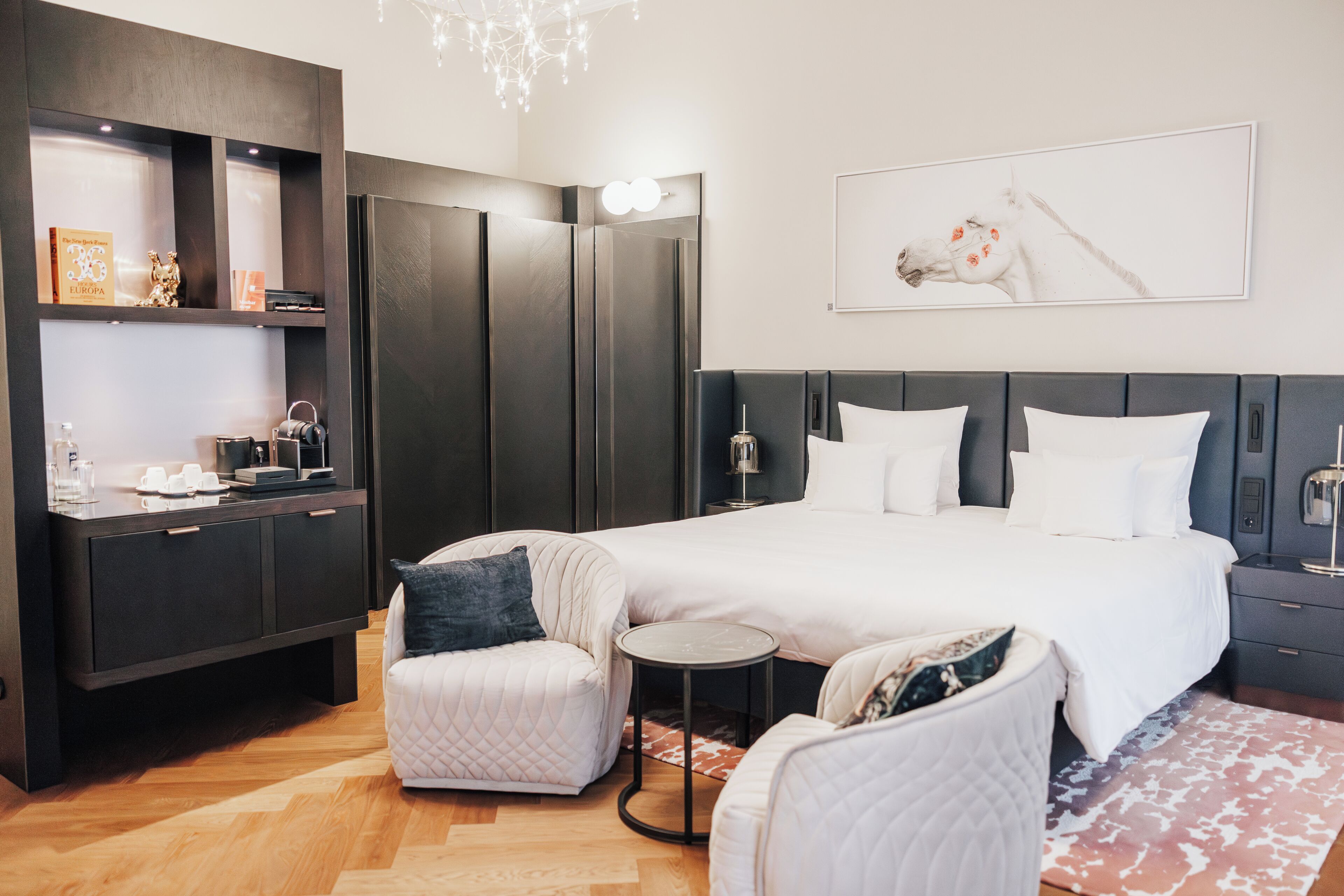 A modern hotel room with a large bed, a seating area with a small table, a wardrobe, and a wall-mounted artwork.