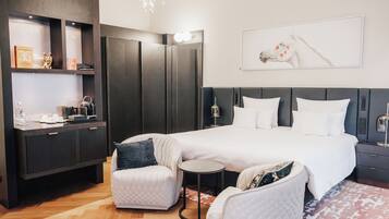 A modern hotel room with a large bed, a seating area with a small table, a wardrobe, and a wall-mounted artwork.