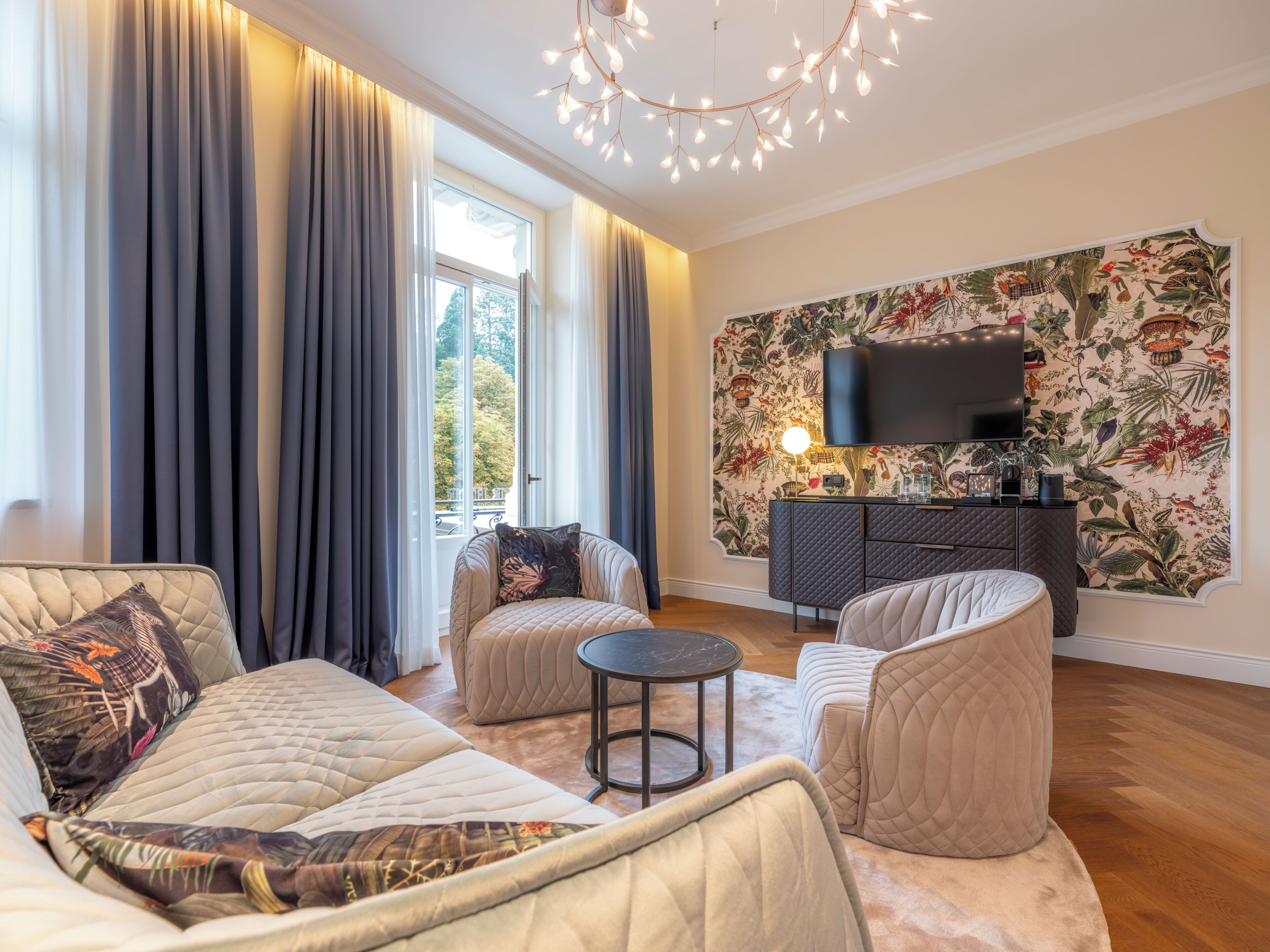 Un salon moderne avec un canapé, des fauteuils et une petite table ronde. Un téléviseur est fixé au mur et il y a une fenêtre avec des rideaux.