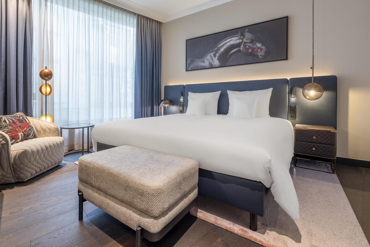 Une chambre d’hôtel moderne avec un grand lit, une petite table de chevet, une chaise et une grande fenêtre avec des rideaux.